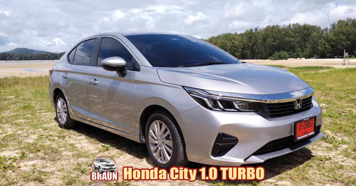 taken in the holiday destination of phuket honda city turbo in silver showing two of its 5 doors parked on sand and grass area before a secluded beach in chalong bay with a wooden structure, forest trees and mountains against a cloudy sky with the tide out